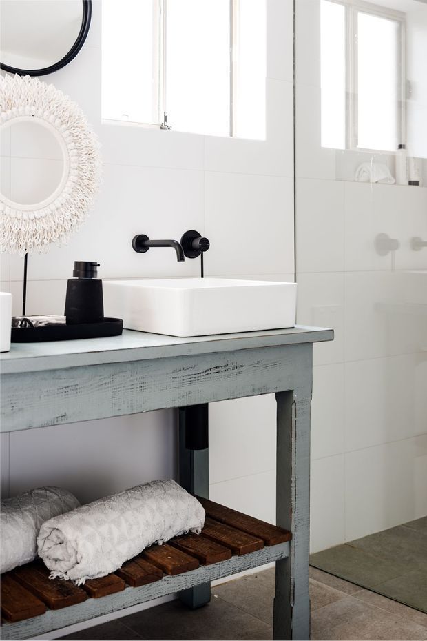 Bright bathroom featuring a white vessel basin on a distressed pale blue-grey timber vanity, matte black wall-mounted tapware, white tiled walls, a frameless glass shower screen, and a decorative woven circular mirror. Folded towels and timber slats occupy the open lower shelf.