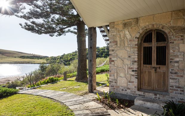 Sunny exterior view of a rustic stone building beside a tranquil lake or inlet. The facade features rough stone masonry, an arched timber-framed door, timber posts, and a covered verandah with a pale lined soffit. A weathered timber walkway crosses the lawn toward the water, with mature trees and rolling countryside beyond.