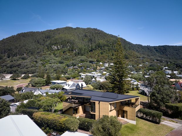 Aerial exterior view of a contemporary single-storey home with a low-pitched dark roof, warm timber-clad walls, extensive glazing, and a compact angular form. The house sits within a leafy suburban neighbourhood, with a densely forested mountain or hill rising behind it.