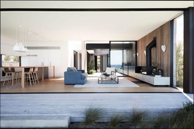 Bright open-plan contemporary living space with a dining area and kitchen to the left, a blue sofa and low coffee table in the centre, and a timber-clad feature wall with a large black fireplace and white floating media console to the right. Full-height glazing opens the room toward a coastal-looking exterior, while a broad pale timber deck and planted grasses sit in the foreground.