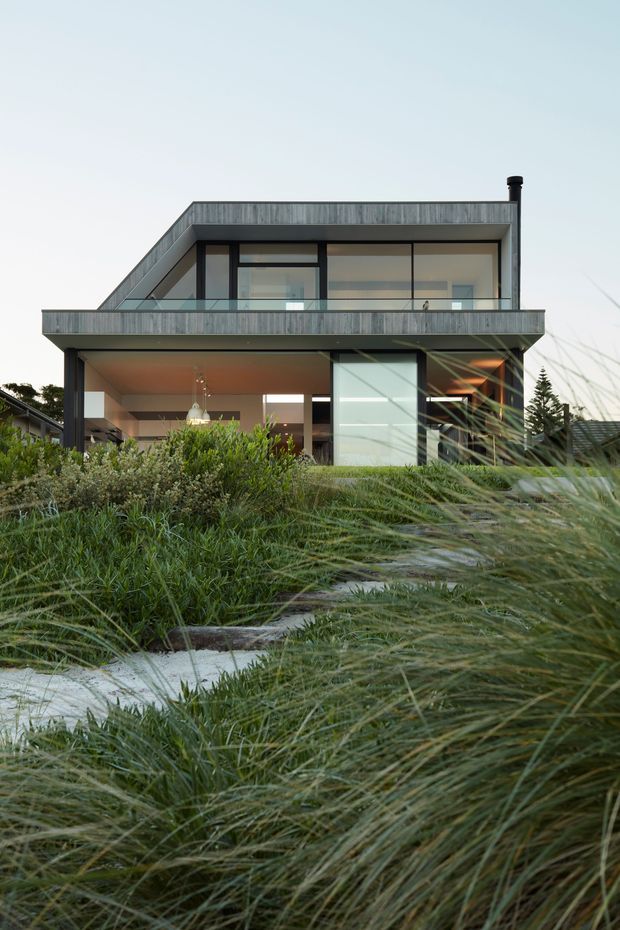 Exterior view of a contemporary two-storey house with a flat roof, extensive glazing, projecting upper-level balcony, dark structural framing, and weathered grey cladding. The home is set behind dense ornamental grasses and a pale stepping-stone path, with warm interior lighting visible through the glazing.