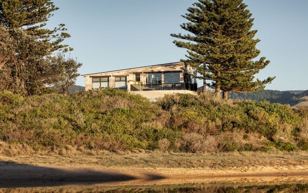 A low, modern single-storey house set on a scrub-covered coastal or elevated site. The building has a long horizontal form, expansive dark-framed glazing, pale masonry or concrete walls, and a flat or very low-pitched roof. Mature wind-shaped conifer trees and distant rugged terrain surround the residence.