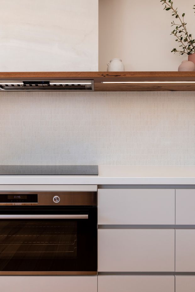 Close-up view of a refined contemporary kitchen with integrated cabinetry, a built-in stainless-steel and black oven, a slim induction cooktop, pale textured tiled splashback, and a warm timber open shelf with minimalist ceramic vessels and a vase. The composition emphasizes clean horizontal lines, subtle neutral tones, and understated joinery detailing.