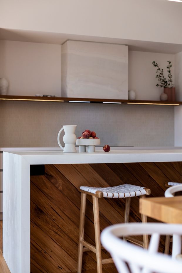 A close-up view of a refined contemporary kitchen featuring a white waterfall-edge island with a dark, diagonally laid timber feature panel on the front. Pale cabinetry and wall surfaces are paired with a long slim timber shelf, integrated warm lighting, a subtle textured splashback, and minimal ceramic decor. A woven timber bar stool and partial dining furniture are visible in the foreground.