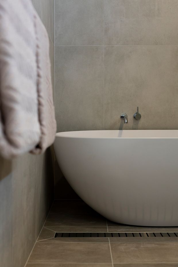 Close-up view of a contemporary bathroom featuring a freestanding white bathtub against large beige stone-look wall tiles. A wall-mounted chrome tap and control are visible above the tub, with matching beige tiled flooring and a linear floor drain. A soft pale towel hangs in the foreground.