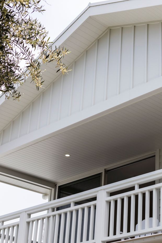 Close-up exterior view of a bright white gabled residential facade, showing vertical wall cladding, a pitched gable roof, a covered balcony or veranda with white balustrades, a lined soffit with recessed lighting, and dark-framed glazing. Tree branches partially frame the upper left.
