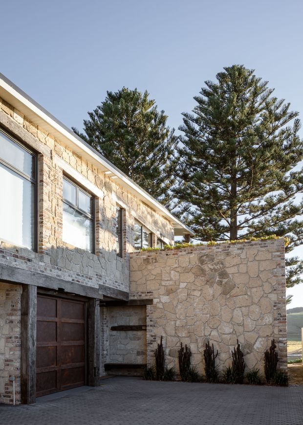 Exterior view of a low-slung stone and brick residential building with large windows, a dark timber garage door, a paved driveway, and a substantial stone courtyard wall. Tall conifer trees rise behind the building, with a rural or coastal landscape faintly visible beyond.