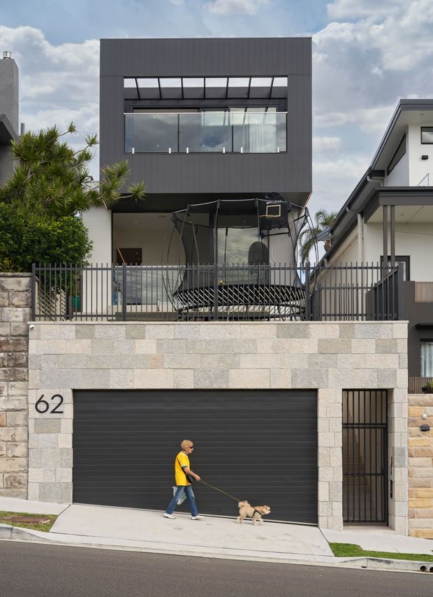 Street-facing exterior view of a contemporary multi-level urban residence. The facade features a dark charcoal upper volume with vertical cladding, large glazed windows and a projecting roof or balcony edge, set above a pale stone-clad garage level. A fenced elevated terrace contains a trampoline, and a separate side gate provides access. Neighbouring modern homes, landscaping and a pedestrian with a dog are visible in the surroundings.
