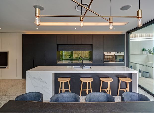 Contemporary open-plan kitchen and dining area dominated by a large white stone island with dark integrated cabinetry, four timber stools, and a dark timber dining table in the foreground. The kitchen includes a window splashback, integrated double ovens, black tapware, and expansive glazing to the right. A linear geometric pendant light hangs beneath a recessed ceiling feature, with neutral terrazzo-like flooring and dark blue upholstered dining chairs.