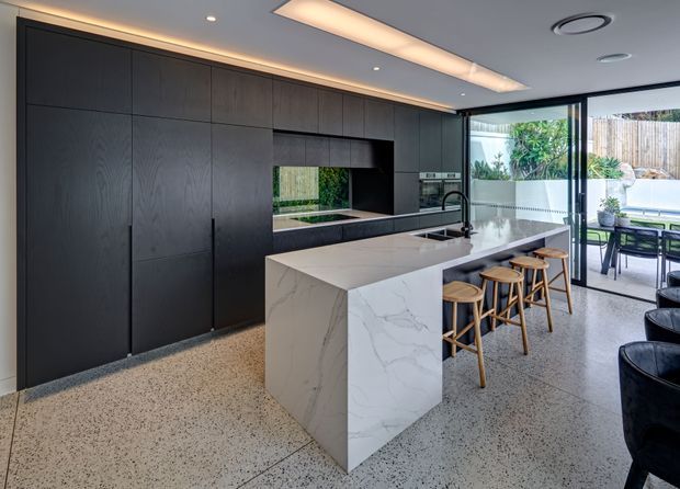 Contemporary kitchen with a full-height dark timber-toned cabinetry wall, integrated appliances, a large white veined stone island with waterfall edge, and timber stools. Terrazzo flooring, recessed and linear ceiling lighting, and extensive black-framed glazing connect the kitchen to an outdoor dining area.