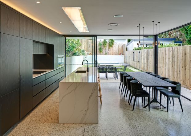Contemporary open-plan kitchen and dining area opening onto a landscaped outdoor courtyard. The kitchen features dark timber-look full-height cabinetry, an integrated dark splashback, a pale stone island with waterfall end, and a black tap and sink. A long dark dining table with upholstered chairs sits beneath slim pendant lights, while extensive glazing and sliding doors connect the space to the outdoor area. Polished terrazzo-style flooring, recessed lighting, and concealed warm cabinet lighting create a refined indoor-outdoor setting.