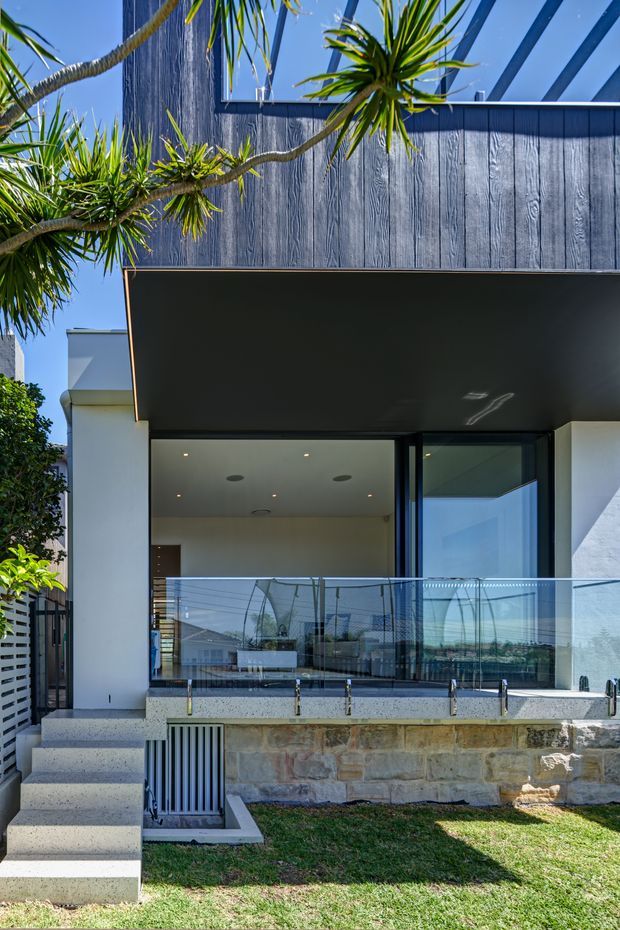 A contemporary multi-level residential facade viewed from the garden, featuring dark vertical timber-style cladding, white rendered walls, a deep flat-roofed canopy, extensive glass doors and balustrading, and a raised terrace supported by a stone retaining wall. Tropical cordyline planting frames the upper facade, while concrete steps lead up to the outdoor living area.