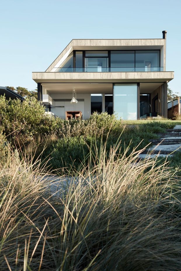 Exterior view of a contemporary two-storey residence with a flat roof, broad cantilevered balcony, extensive black-framed glazing, and pale timber-clad upper-level walls. The ground floor opens toward a landscaped coastal-style garden with ornamental grasses and a stepped path. A covered outdoor living area and interior dining space are visible through the glazing.