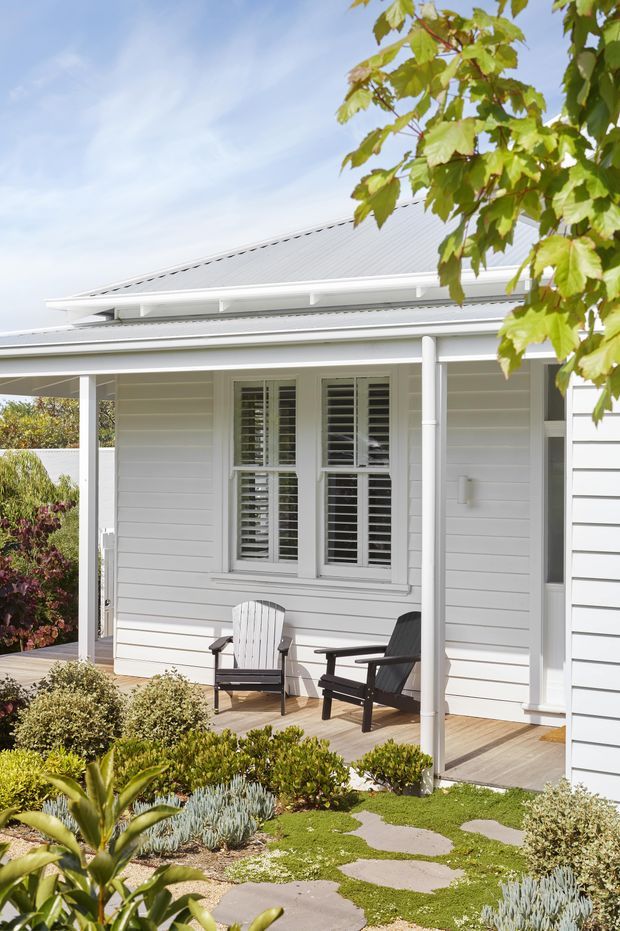 Bright exterior view of a single-storey light-coloured weatherboard home with a covered front verandah, corrugated pitched roof, white-framed shuttered windows, timber decking, outdoor chairs, and a landscaped garden with stepping stones and low planting.