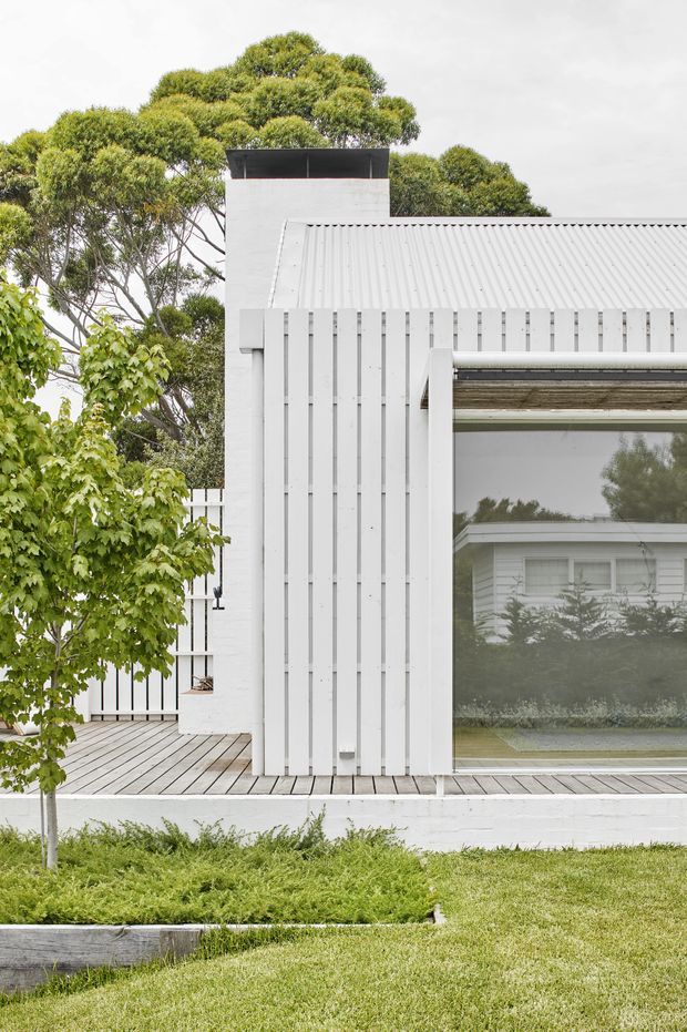 Exterior view of a light-painted, contemporary house with a corrugated metal pitched roof, white vertical timber-style cladding, large glazed opening, brick chimney, and a timber deck set within a landscaped garden.