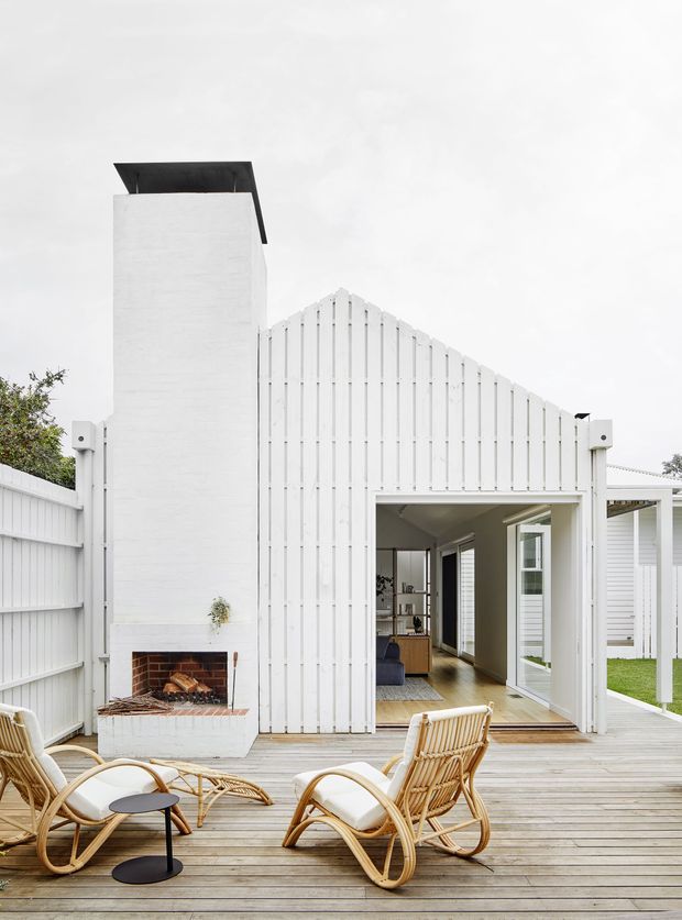 Bright exterior view of a white weatherboard-clad house opening onto a timber deck. The facade features a steep gabled volume, a tall chimney with an outdoor brick fireplace, and large glazed doors leading into a light-filled interior. Wicker lounge chairs and a small black table sit on the deck, with a covered outdoor area visible to the right.
