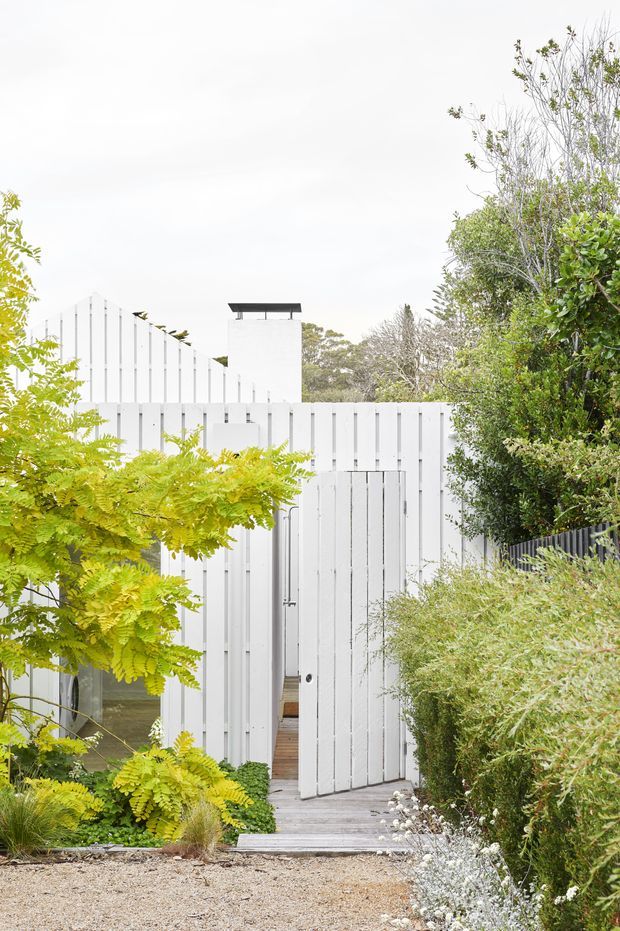 Exterior view of a contemporary white-clad house or garden structure, approached via a timber walkway through dense landscaped planting. The facade features vertical board cladding, a tall white chimney with a dark cap, and a partially open vertical-board gate or door. The scene has a minimalist architectural composition softened by lush greenery and a pale gravel foreground.