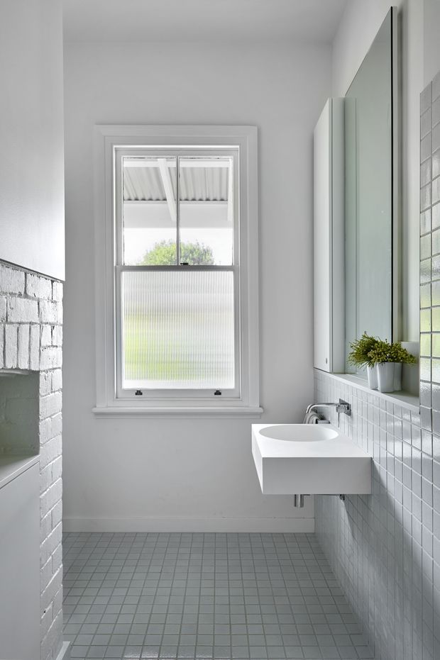 Bright, compact contemporary bathroom with a wall-mounted white vanity and integrated basin, large mirrored cabinet, pale square tiled walls and floor, and a white-painted brick partition enclosing the bath or shower area. A framed window provides natural light, with a small potted plant on the vanity.