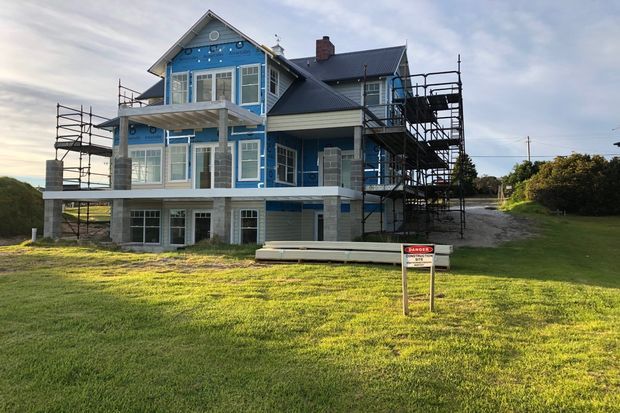A multi-storey residential house under construction, viewed from the front. The building has blue weatherboard-style exterior cladding, white trim, a dark pitched roof, multiple balconies, extensive scaffolding, and exposed construction wrapping. It sits on a grassy suburban or semi-rural site with a driveway to the side.
