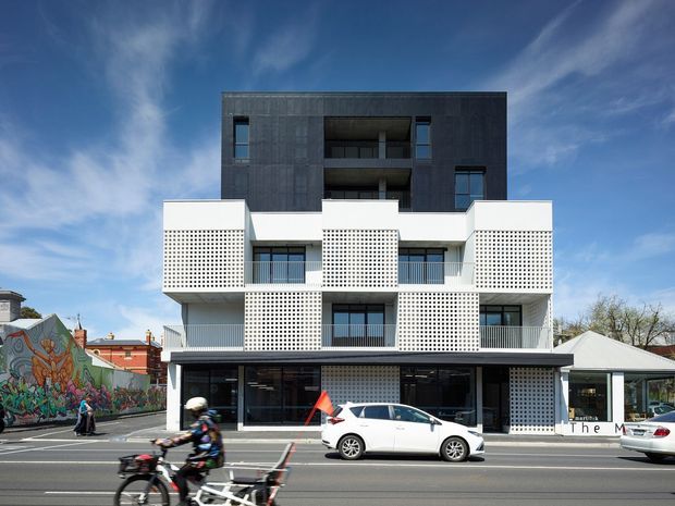Street-facing multi-storey apartment building with a contemporary facade combining dark upper-level cladding, white rendered bands, and extensive perforated breeze-block screens around balconies. The ground floor has dark commercial-style glazing and recessed entrances, while parked cars, cyclists, neighbouring buildings, and a mural-lined street appear in the foreground and surroundings.