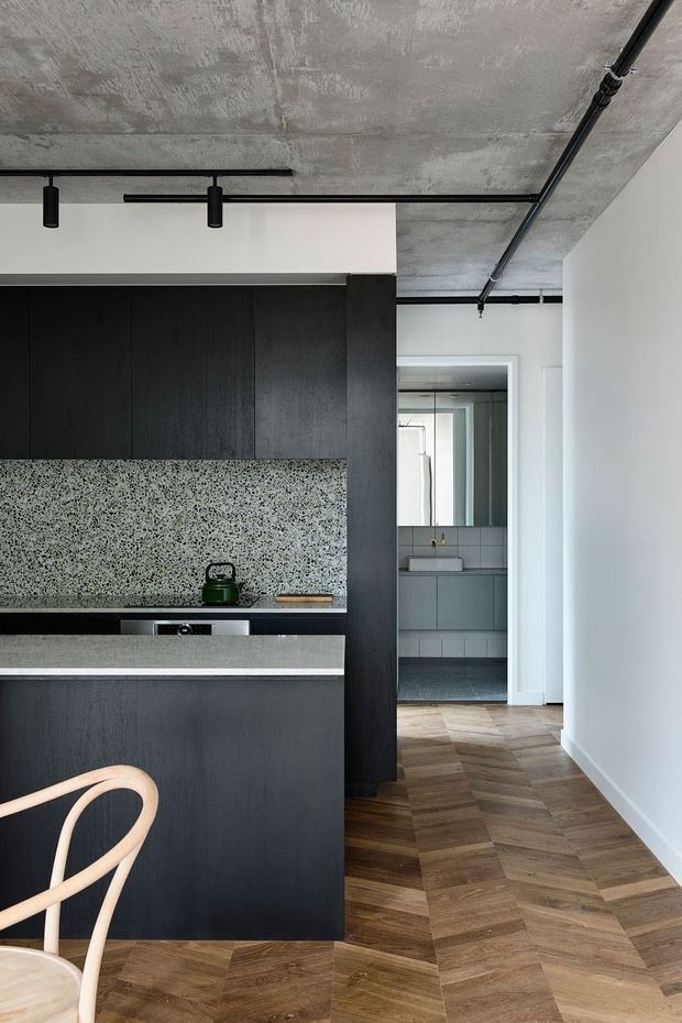 A compact contemporary kitchen with matte black full-height cabinetry, a speckled terrazzo-style backsplash, and a pale stone or engineered-stone benchtop. The kitchen opens toward a tiled bathroom visible in the background. Herringbone timber flooring, exposed raw concrete ceilings, and black surface-mounted lighting and conduit create an industrial character, while white walls keep the space bright and minimal.