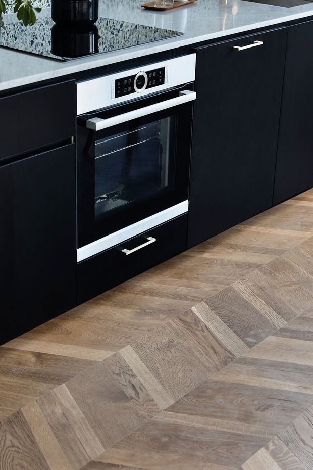 Close-up view of a contemporary kitchen featuring matte black cabinetry, a built-in stainless-steel-trimmed oven, pale stone benchtops, and a terrazzo-style backsplash. The floor is finished in a prominent light-to-mid wood herringbone pattern, with minimal black and metallic hardware.