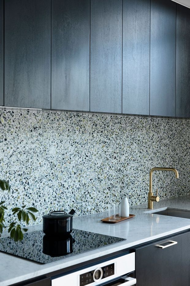 Close-up view of a sophisticated contemporary kitchen featuring dark vertically panelled upper cabinetry, a pale terrazzo-style full-height splashback, a light stone benchtop, black induction cooktop, integrated oven, and a brushed brass gooseneck tap. The palette is cool and refined with strong tonal contrast and a small amount of greenery and countertop accessories.