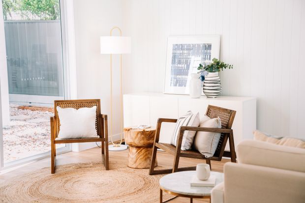 Bright, airy living room with white vertical wall panelling, large glazing, a natural woven rug, warm timber lounge chairs, a pale upholstered sofa, a round marble coffee table, and a slim brass floor lamp. Minimal artwork and a potted plant add restrained styling, while a gravel courtyard is visible outside.