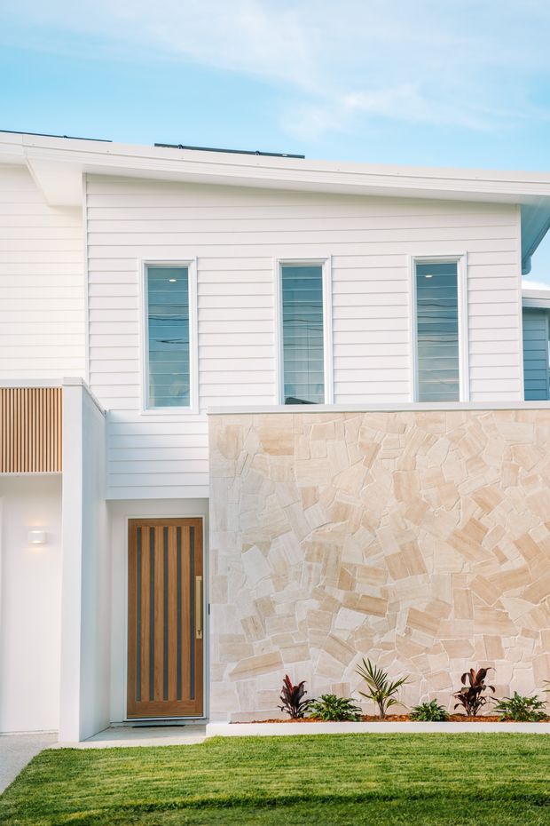 Exterior view of a contemporary two-storey residence with white horizontal weatherboard cladding, a broad low-pitched roofline, tall narrow windows, and a timber front door. A prominent pale irregular stone feature wall projects across the facade, with a narrow planted garden bed and manicured lawn in front. The image has a bright, clean coastal feel.