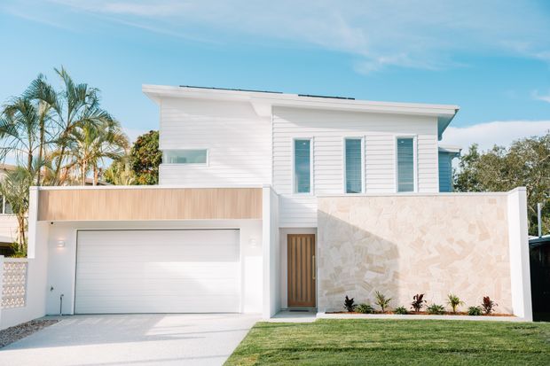 Bright contemporary two-storey residential facade with white horizontal weatherboard cladding, a low-pitched roof, tall narrow windows, a white garage, timber-toned garage surround, timber entry door, and a pale stone feature wall. A neat lawn, driveway, and tropical planting frame the front elevation.