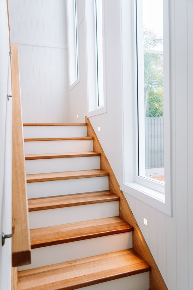 Bright interior stairway with warm timber treads and handrail, white painted vertical wall panelling, and tall white-framed windows bringing in abundant natural light. The stair rises through a narrow, clean-lined space with a view of exterior planting through the glazing.