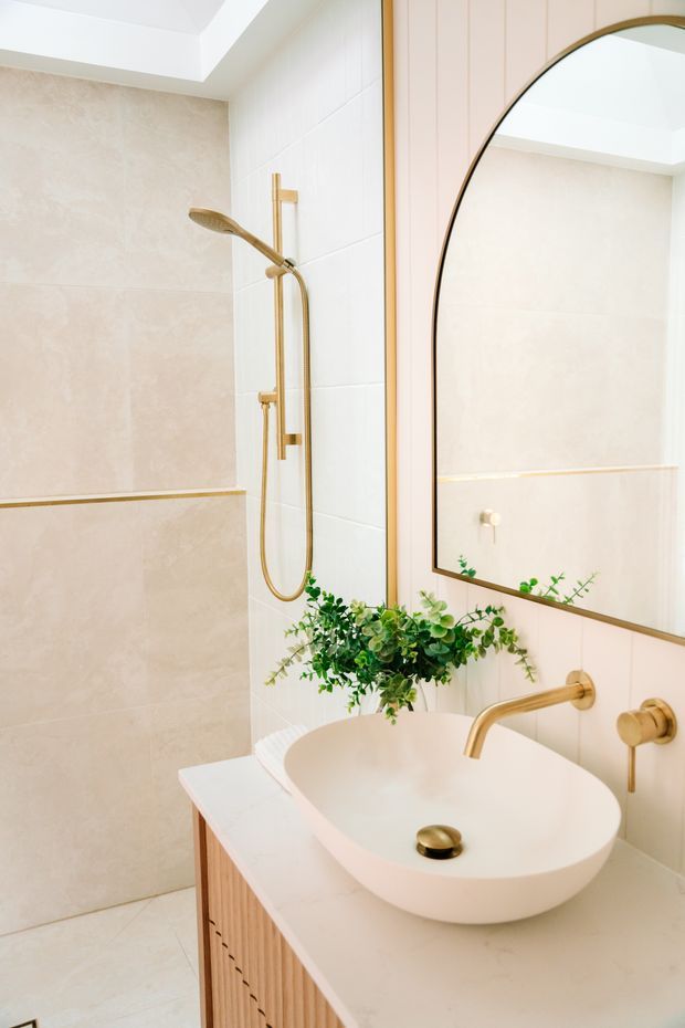 A bright, refined bathroom featuring a walk-in shower with large beige wall tiles, a white tiled shower wall, brushed gold shower fittings, and a floating vanity with a white vessel basin. The vanity has light timber fluted cabinetry and a pale stone or solid-surface countertop, accompanied by a large rounded mirror with a slim dark-gold frame. Greenery adds a natural accent, and a skylight or recessed ceiling opening provides abundant light.