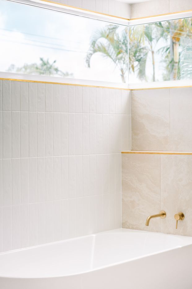 Bright bathroom featuring a white freestanding or built-in bathtub beneath a large corner window. The walls combine vertically grooved white tiles with pale beige stone-look tiles, accented by slim gold trim lines and brushed gold bath fittings. Tropical palms are visible outside the glazing.