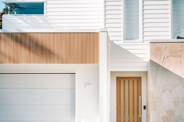 Close-up exterior view of a contemporary multi-level residential facade. The building features white horizontal weatherboard cladding, a warm timber slat balcony privacy screen, white-framed glazing, a sectional garage door, and a timber front door. A light natural-stone boundary wall occupies the right side.