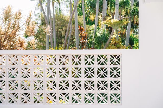 A white decorative breeze-block screen with repeating geometric floral cutouts, set within a clean white wall. Lush tropical foliage and slender palm-like trees are visible behind the screen, creating a bright indoor-outdoor architectural feature.