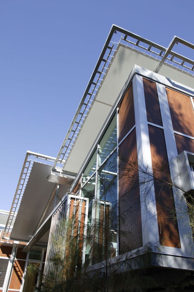 Low-angle exterior view of a multi-storey contemporary building with projecting flat roof planes, prominent glazed façades, white structural framing, and warm timber-clad vertical wall panels. Slim metal roof-edge railings and balconies add strong horizontal and linear detailing, with landscaped planting at the base.