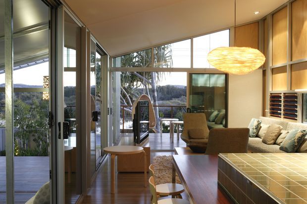 A warm, contemporary open-plan living space with extensive floor-to-ceiling glazing opening toward a raised outdoor deck and leafy landscape. The room includes a large tiled kitchen island in the foreground, timber flooring and built-in timber wall cabinetry, a lounge area with a sofa and armchairs, and a sculptural woven pendant light. The glazing, timber finishes and open connection to the deck create an airy indoor-outdoor setting.
