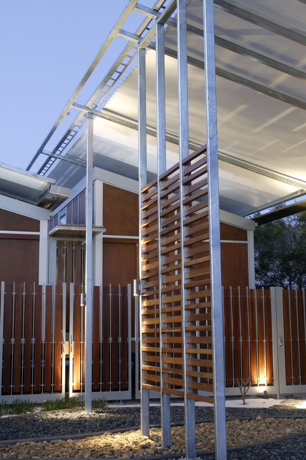 Exterior architectural photograph of a contemporary building with a dramatic elevated flat roof and exposed galvanized steel columns and framing. Timber-clad wall panels and vertical timber fencing are paired with a prominent slatted timber privacy screen. The building sits above a gravel-covered landscaped forecourt with integrated ground lighting, viewed against a clear sky.