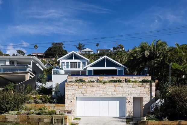 Street-facing view of a coastal suburban residential property on a sloping site. The foreground features a pale stone-clad garage and retaining wall, with a blue-and-white gabled house behind it, landscaped terraces, palm trees, and overhead utility wires under a clear blue sky.
