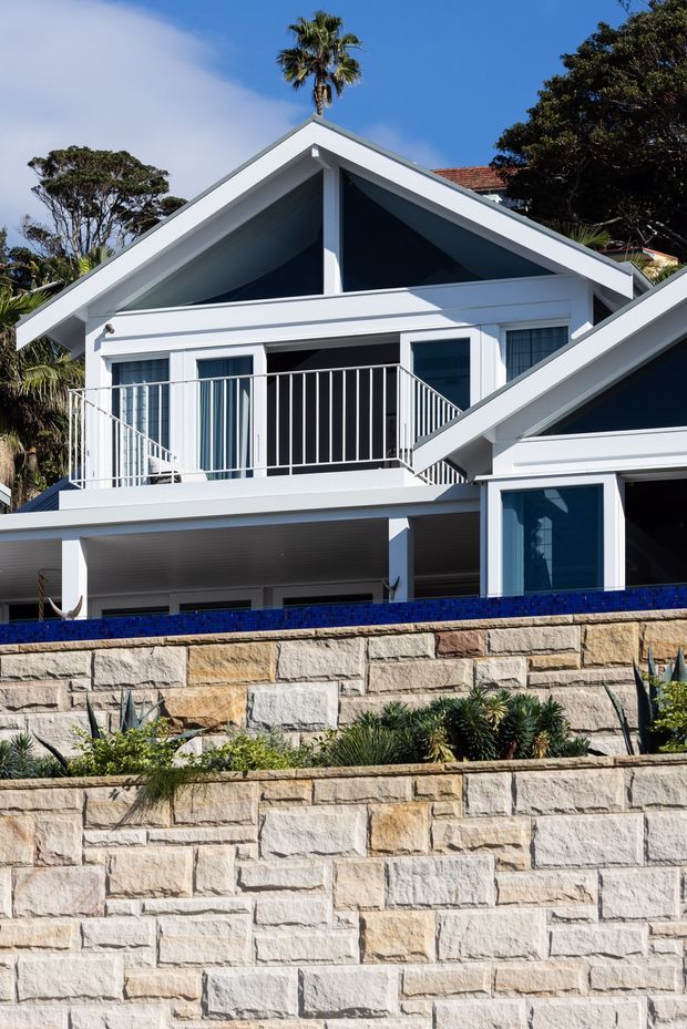 Exterior view of a bright white multi-level coastal-style house with prominent pitched gable roofs, large dark-framed glazing, balcony balustrades, deep eaves, and covered outdoor areas. Terraced sandstone retaining walls and blue-tiled edging occupy the foreground, with palms and established landscaping around the property.