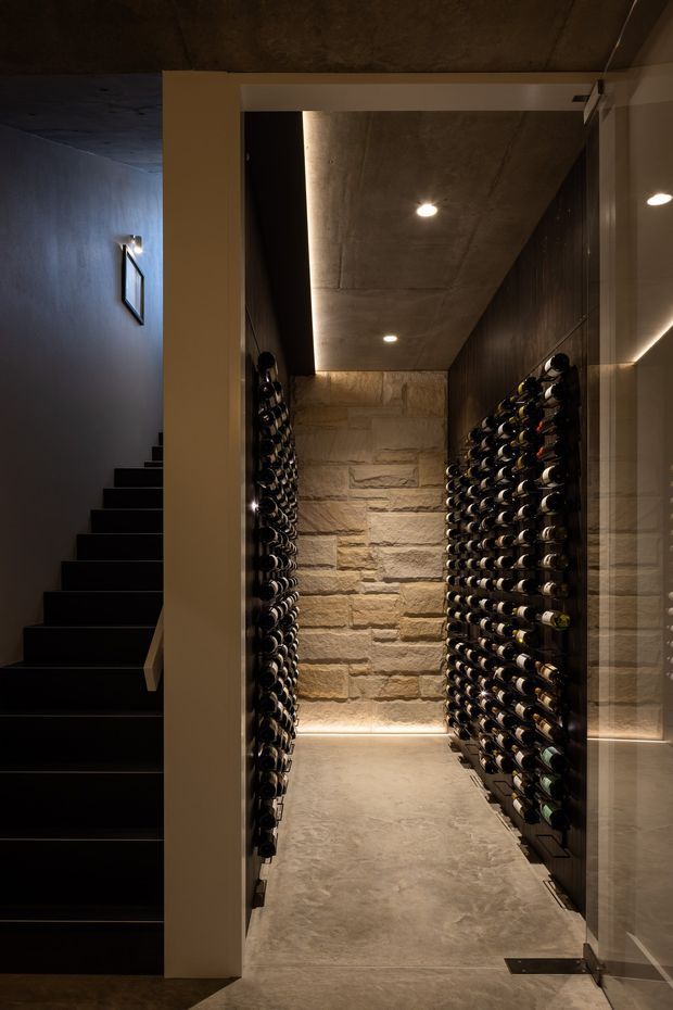 A dark, narrow residential wine cellar viewed through an open glass door, with extensive wall-mounted bottle racks on both sides, a rough stacked-stone feature wall at the rear, and a polished concrete ceiling and floor. A black staircase is visible to the left, while warm recessed and concealed lighting creates an intimate, moody atmosphere.