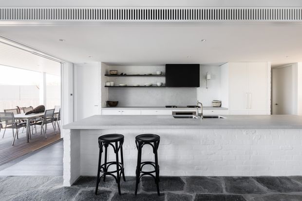 Bright, minimalist open-plan kitchen with a large white-painted brick island, pale stone benchtop, white cabinetry, open shelving, integrated black television, and a stainless-steel sink and tap. Two black stools sit at the island. A dining area and timber-floored outdoor or glazed space are visible to the left. The room combines crisp white finishes with dark slate-like flooring and restrained black accents.