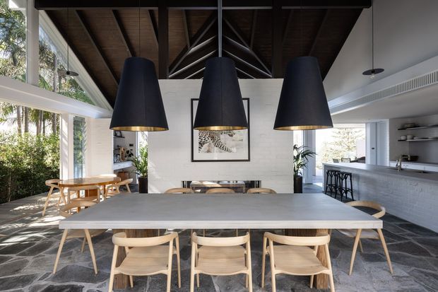 A bright, open-plan dining area centred on a long pale table with light timber dining chairs. Three oversized black pendant lights with brass interiors hang beneath a dramatic dark timber vaulted ceiling with exposed rafters and a central structural truss. Full-height glazing on the left opens to greenery, while a white brick kitchen and open shelving are visible at the right. The space combines natural materials, strong black accents and abundant daylight.