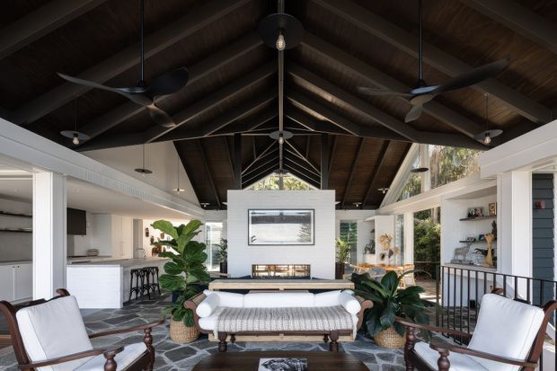 Spacious open-plan living room with a dramatic dark timber vaulted ceiling, exposed rafters, multiple ceiling fans and pendant lights. White walls, extensive glazing, built-in shelving, a central fireplace feature wall, white upholstered seating, patterned stone flooring, and indoor plants create a bright yet warm interior. A kitchen or bar area is visible to the left and a dining or lounge area extends to the right.