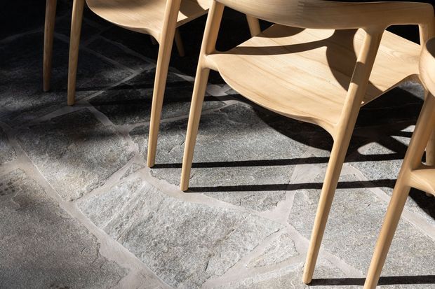 Close-up detail of light timber dining chairs with sculpted seats and tapered legs arranged on a large irregular grey stone floor. Strong sunlight casts crisp shadows across the chairs and flooring; the image focuses on materials, craftsmanship, and texture rather than a complete room.