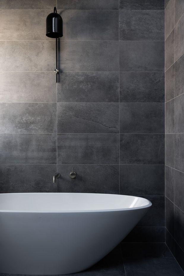 A dark, refined bathroom featuring a freestanding white bathtub against large-format charcoal grey stone-look wall and floor tiles. A black wall-mounted shower fitting and minimalist dark tapware create a strong monochromatic, contemporary aesthetic.