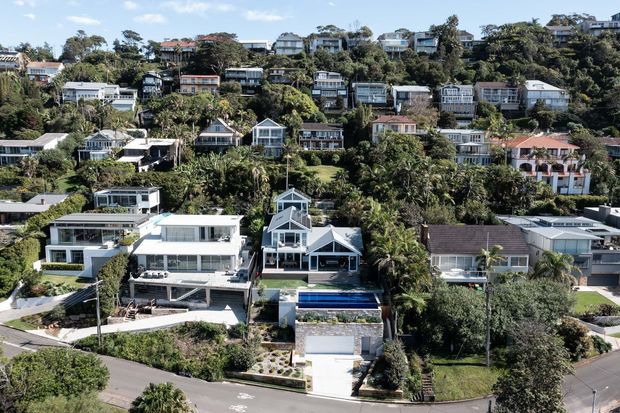 Aerial view of a densely settled, leafy hillside residential neighbourhood with detached homes arranged on steep terraced sites. The foreground includes several contemporary houses, landscaped gardens, driveways, and a narrow swimming pool, while many additional homes and mature trees rise across the slope in the background.