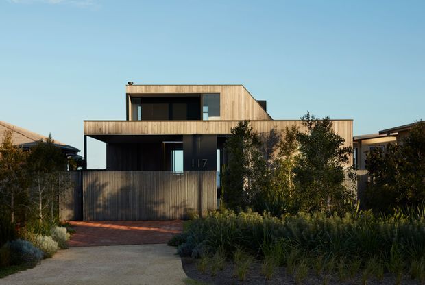 Exterior view of a contemporary multi-level residence with a strong rectilinear form, warm vertical timber cladding, dark-framed glazing, and projecting upper and lower volumes. The house is approached via a paved path and landscaped garden with dense ornamental planting. A neighbouring structure is partially visible at the sides, under a clear sky.