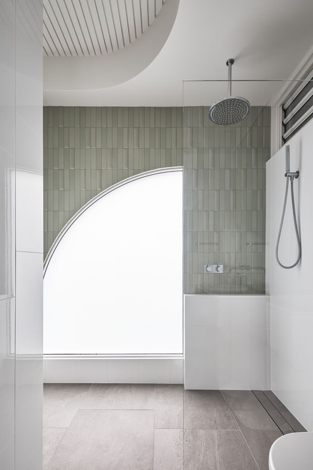 A bright, contemporary bathroom shower area with a curved frosted window, pale sage-green vertical wall tiles, white tiled walls and built-in shower bench, a large round rainfall showerhead, and grey stone-look floor tiles. The room features distinctive curved ceiling and window geometry with clean, minimal detailing.