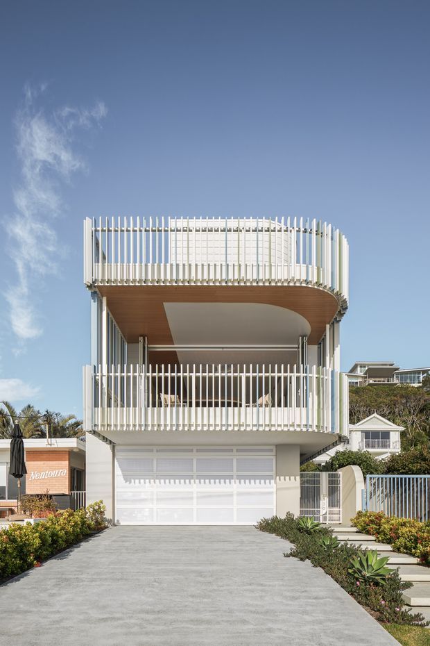 Front exterior view of a contemporary multi-storey coastal residence with a rounded projecting balcony, white vertical screen balustrades, a timber-lined soffit, extensive glazing, and an integrated double garage. A broad concrete driveway and landscaped garden frame the facade.